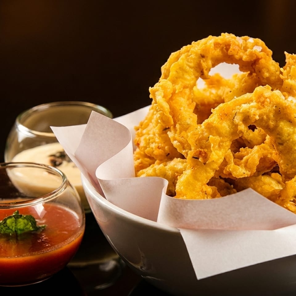 Crispy golden onion rings in a white bowl with two dipping sauces on a dark background.
