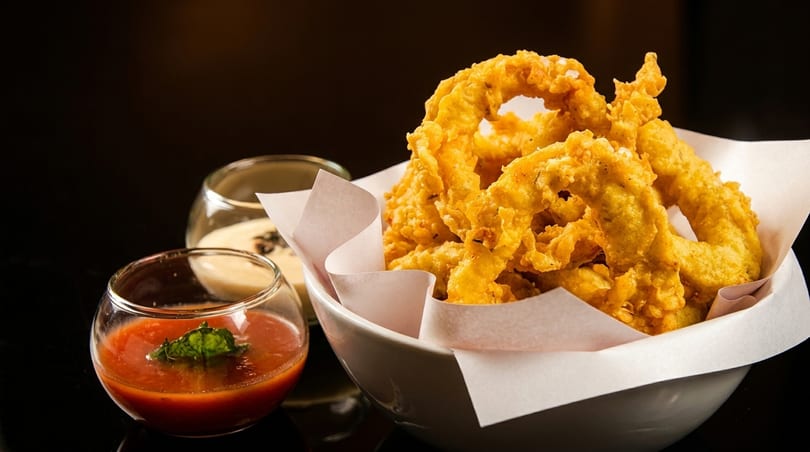Crispy golden onion rings in a white bowl with two dipping sauces on a dark background.