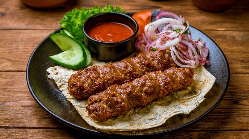 Two grilled meat skewers with sauce, sliced cucumbers, lettuce, and red onions on a black plate.